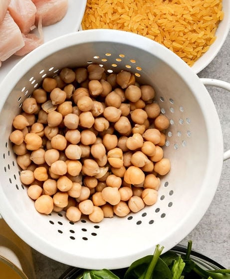 Overhead view white colander holding chickpeas next to other bowls filled with orzo, spinach and chicken breasts.