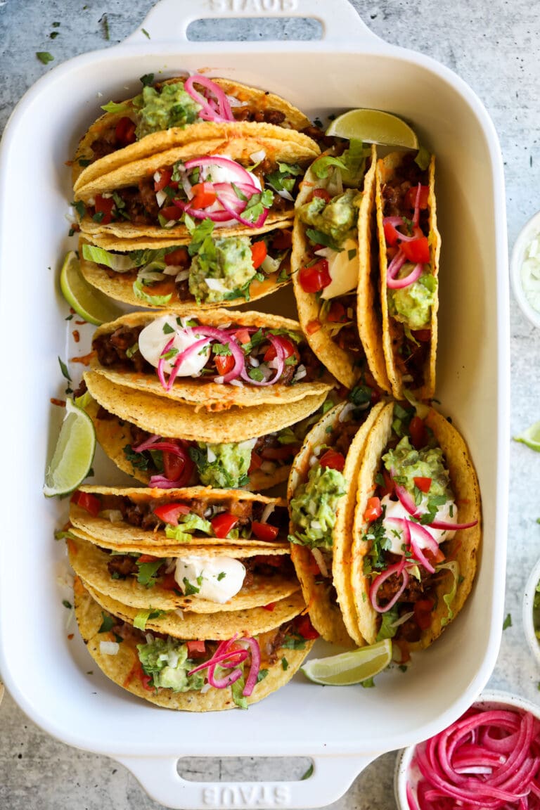 Overhead view white baking dish filled with hard shell tacos filled with beef taco meat, melted cheese and taco toppings