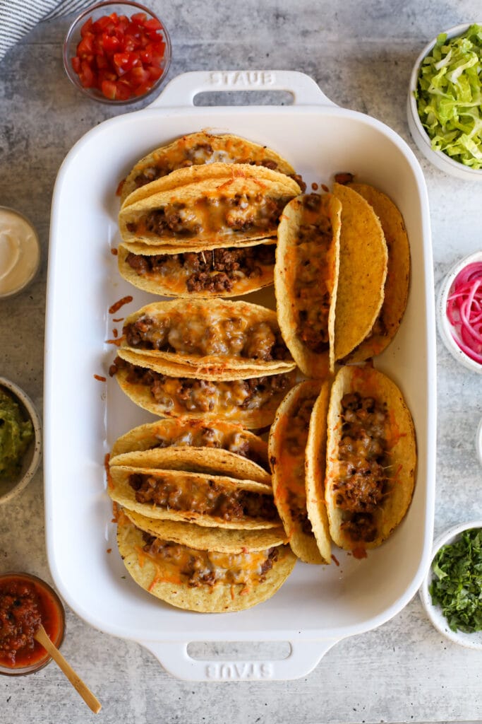 Overhead view of yellow taco shells filled with taco meat and topped with melted cheese in a white baking dish.
