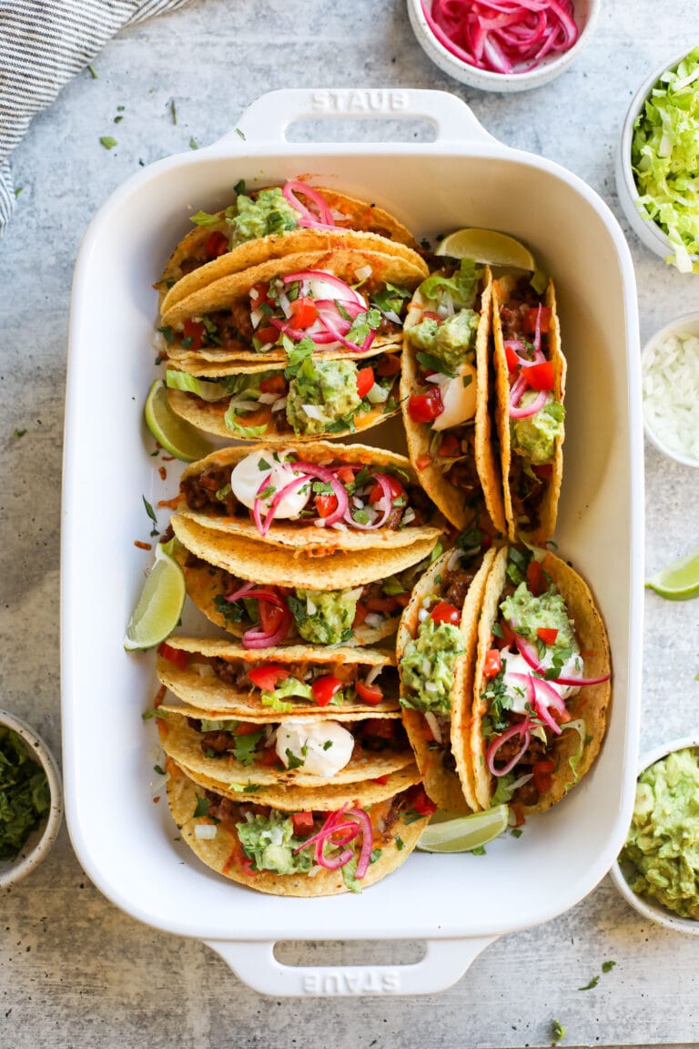 Overhead view of a pan of easy baked tacos topped with pickled onions, guacamole, and lime wedges. 