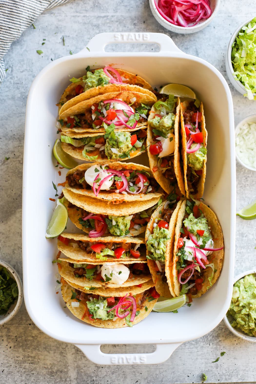 Overhead view of a white baking dish filled with baked tacos and topped with fresh guacamole and garnished with limes.