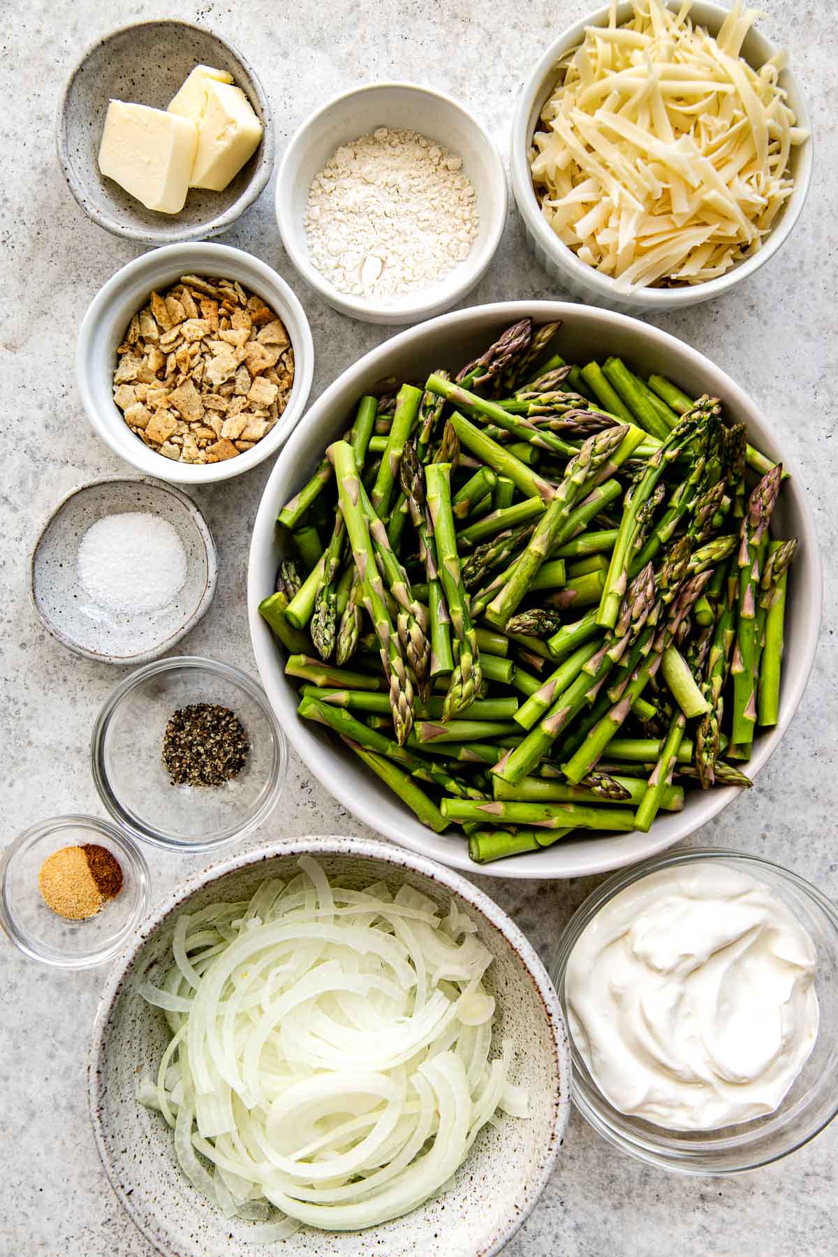 Overhead view of raw ingredients to make an asparagus casserole including fresh asparagus, shredded cheese, crackers, butter, flour, onions and sour cream.