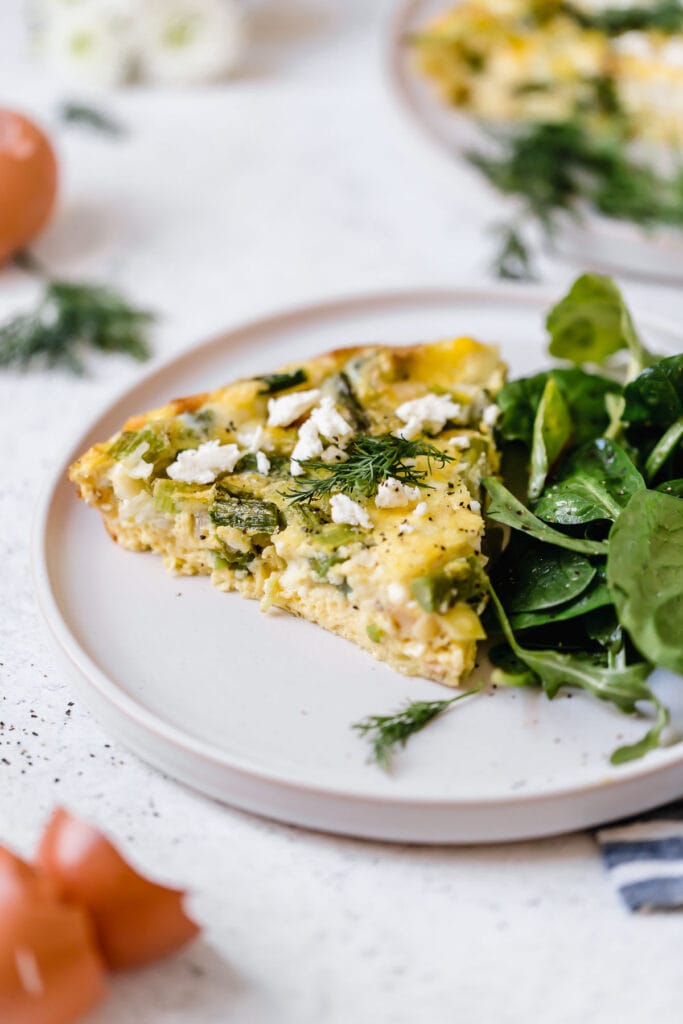 Side view of slice of crustless quiche with asparagus, leek and feta cheese. Served on white plate with spinach salad on side