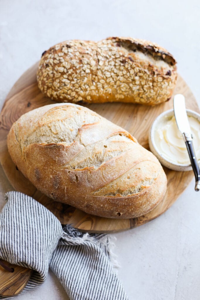 Overhead view of two loaves of sourdough bread on a wooden cutting board next to butter and a butter knife.