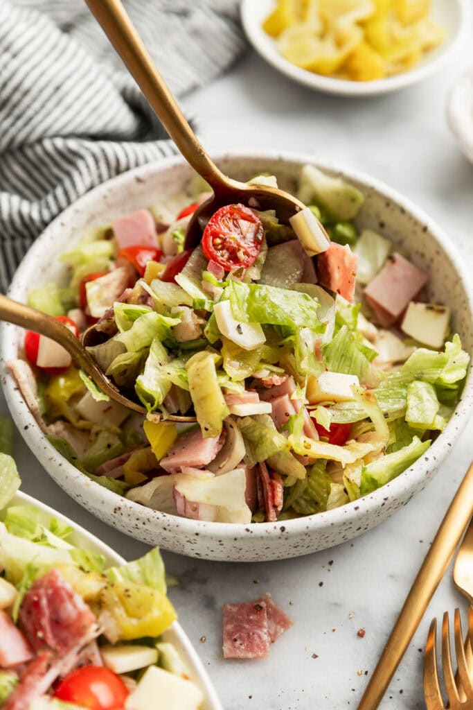 Two spoons mixing and tossing a sub in a tub salad in a white bowl. 