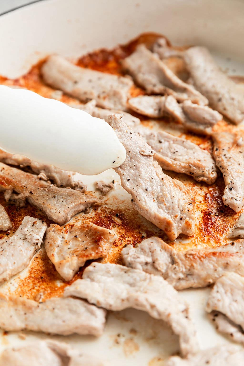 Close up view strips of pork being browned in skillet; tongs turning a strip over to cook on other side
