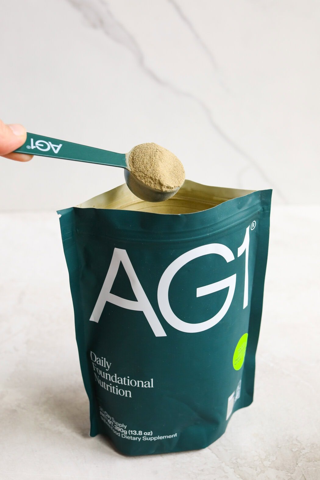 Close up view of a hand using a measuring spoon to scoop out a spoonful of AG1 greens powder out of the packaged bag. 