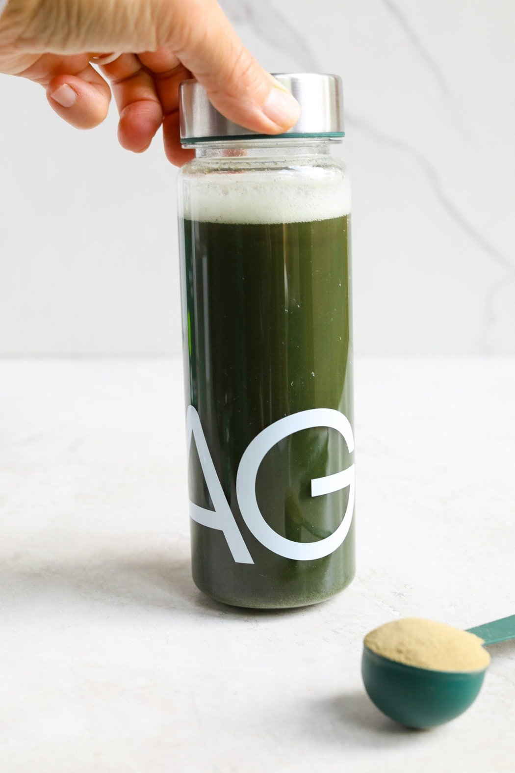Close up view of a clear bottle of AG1 mixed in water to show the green color and frothy foam on top from just shaking. 