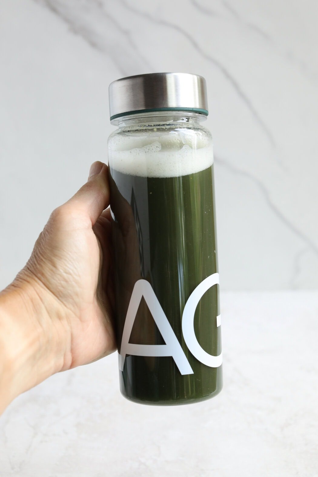 Close up view of a hand holding a clear bottle of mixed AG1 greens showing the green color and foam on top. 