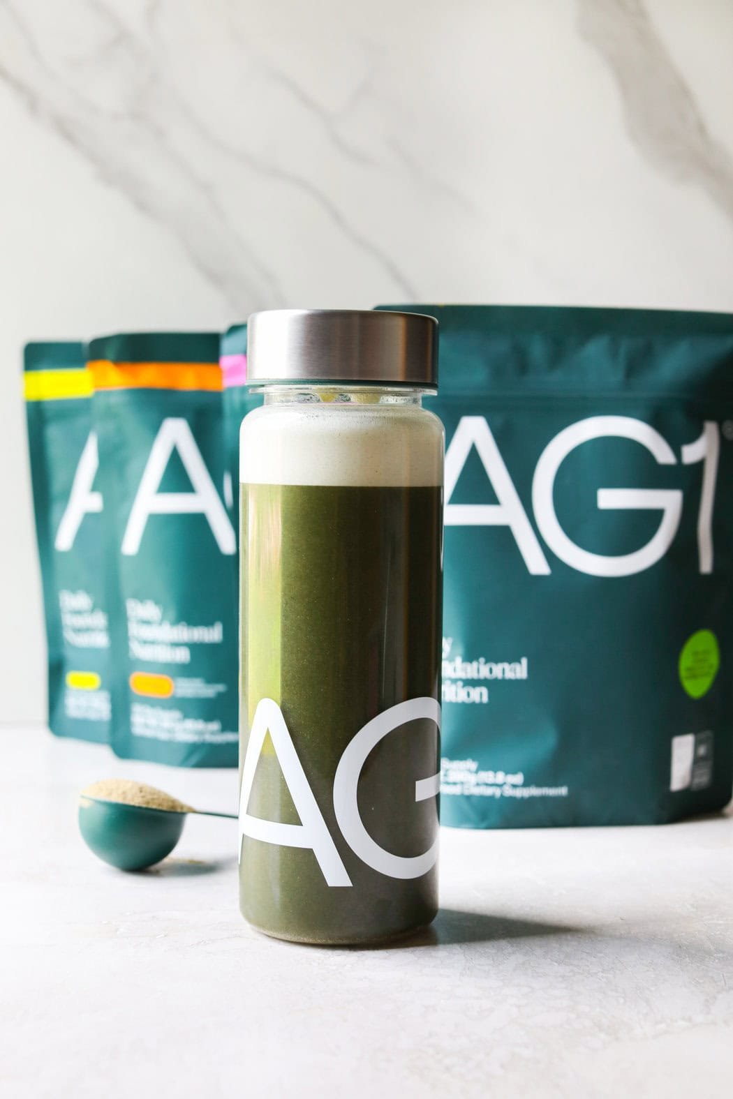 Close up view of a bottle of AG1 greens drink in a clear bottle to show the green color with additional bags behind it. 