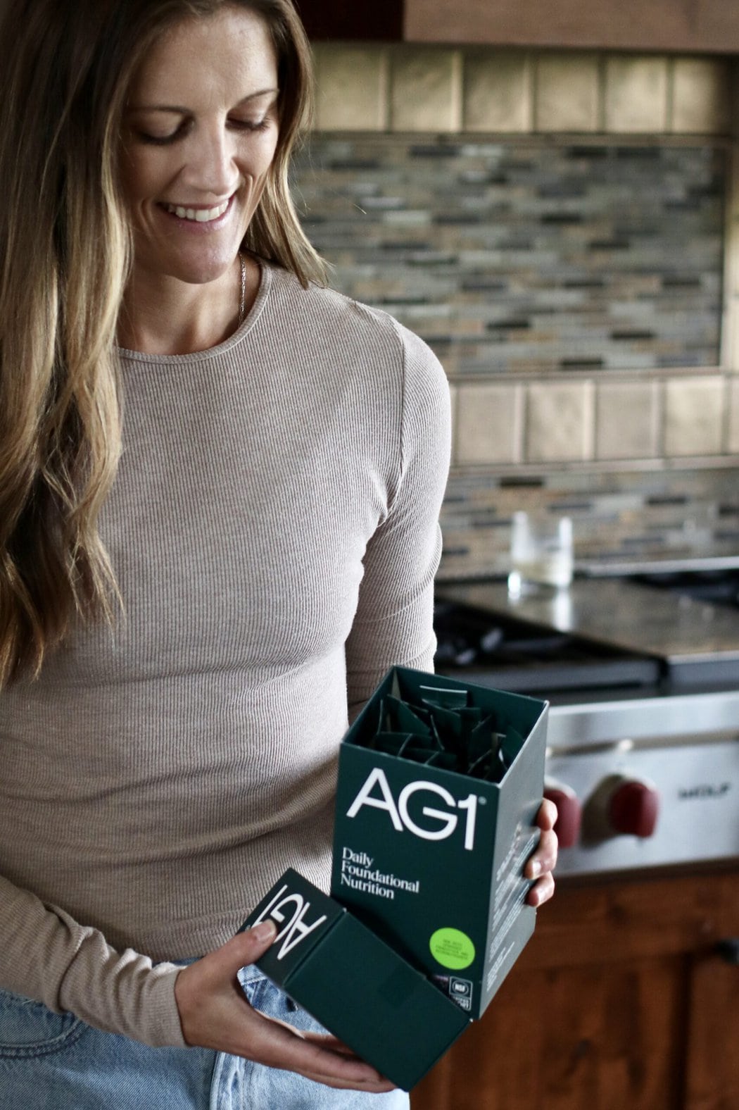 Close up view of a woman in a kitchen opening a box of travel AG1 greens powder sticks. 