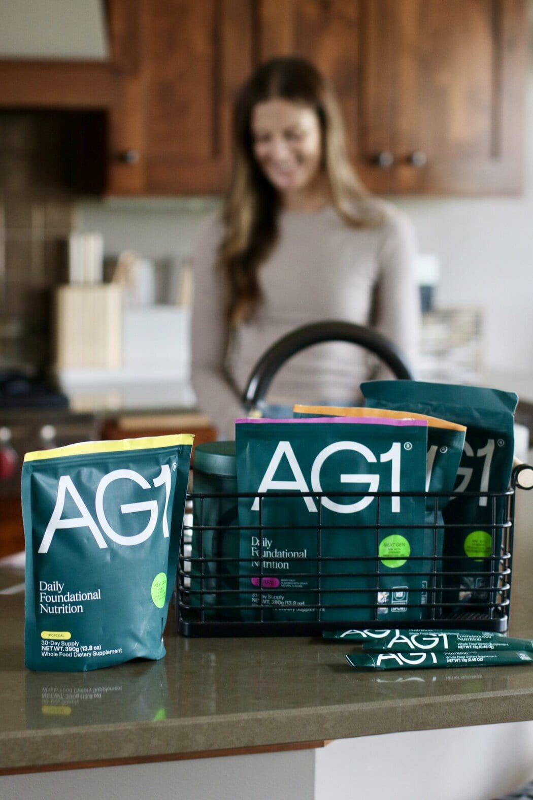 Close up view of a AG1 powdered greens showing packaging and flavor options with a woman in the background of a kitchen. 