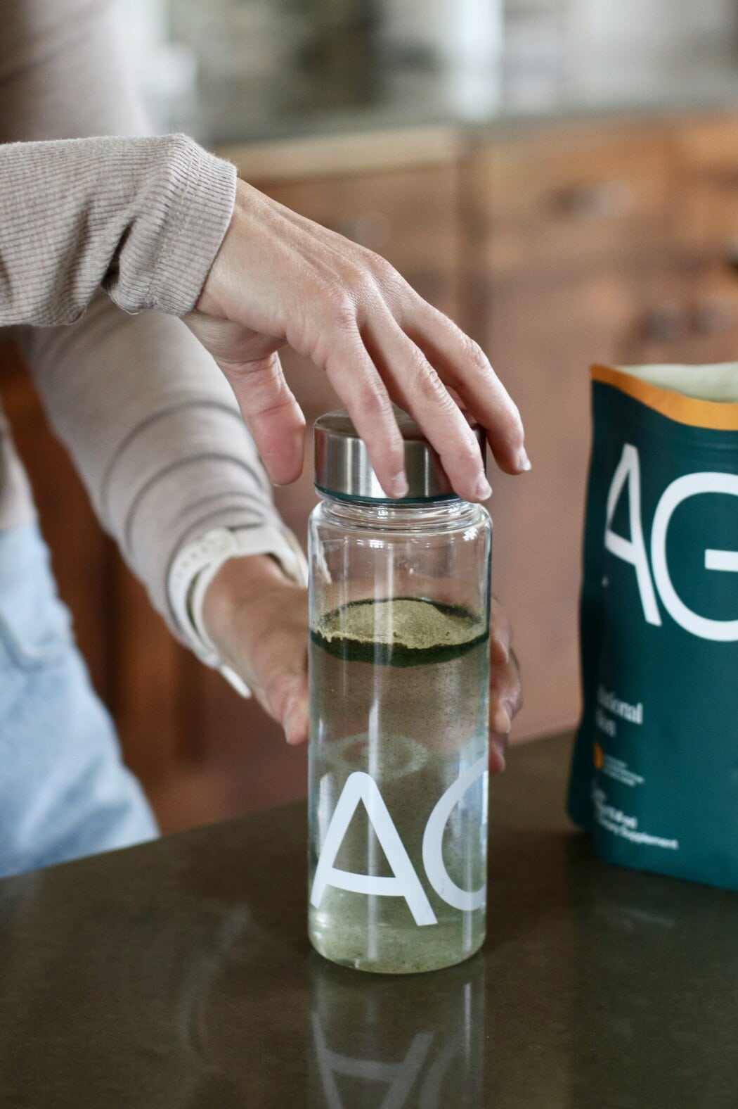 Close up view of a woman at a kitchen counter twisting the cap of a bottle on, showing the AG1 powdered greens on top of the water in the bottle. 