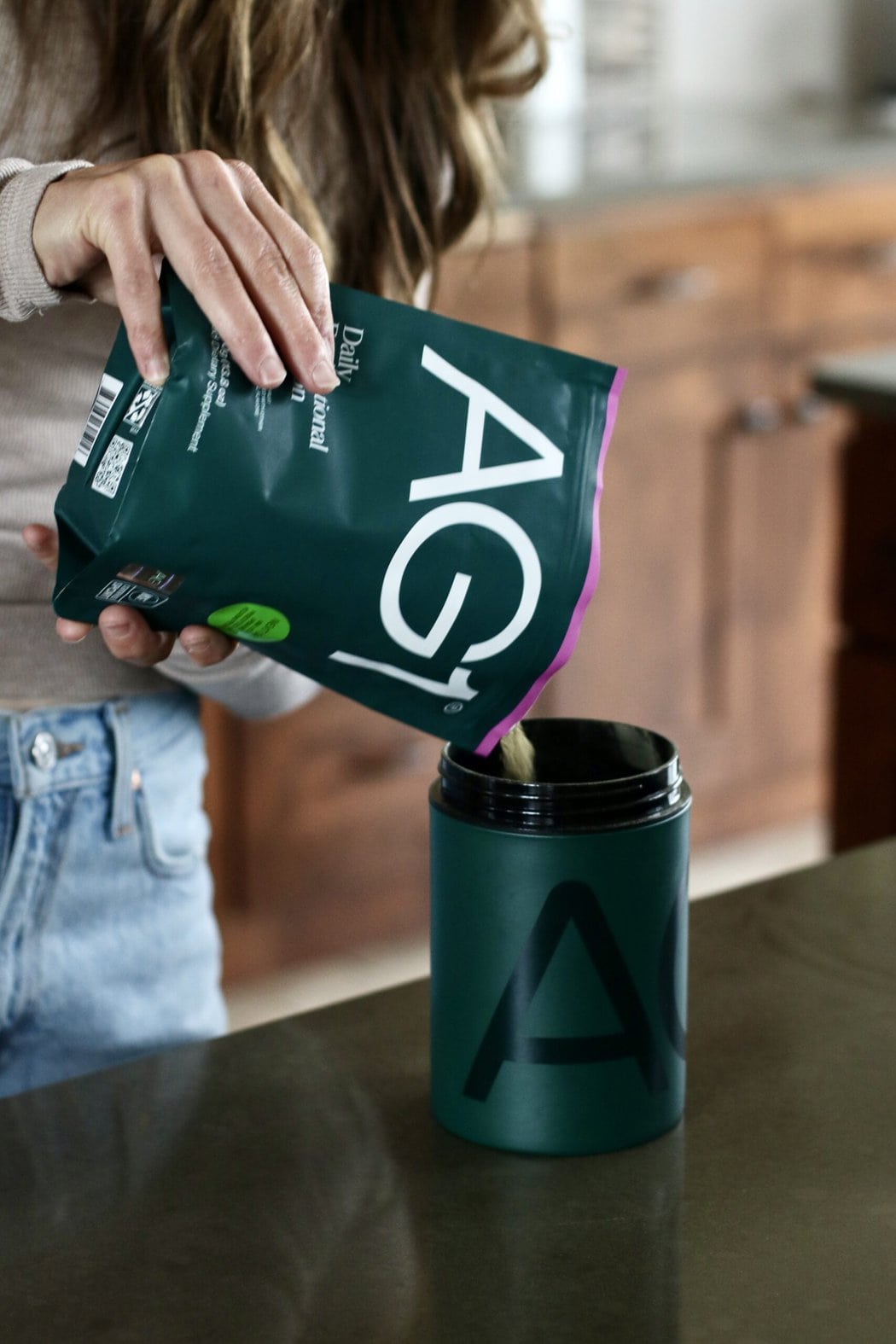 Close up view of a woman at a kitchen counter pouring AG1 greens into a storage canister. 