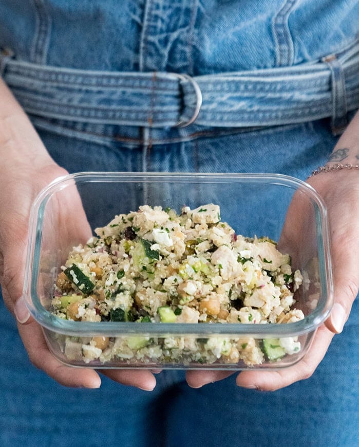 A woman holding a glass meal prep container filled with a quinoa salad topped with fresh mint.