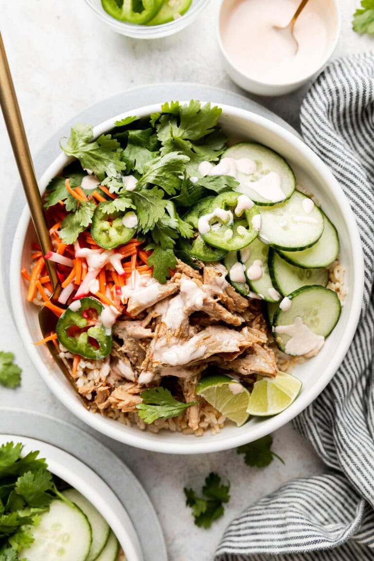 Close up view of a white bowl filled with sliced marinated carrots, cucumbers, fresh cilantro and shredded banh mi pork topped with a creamy dressing and sliced jalapenos.