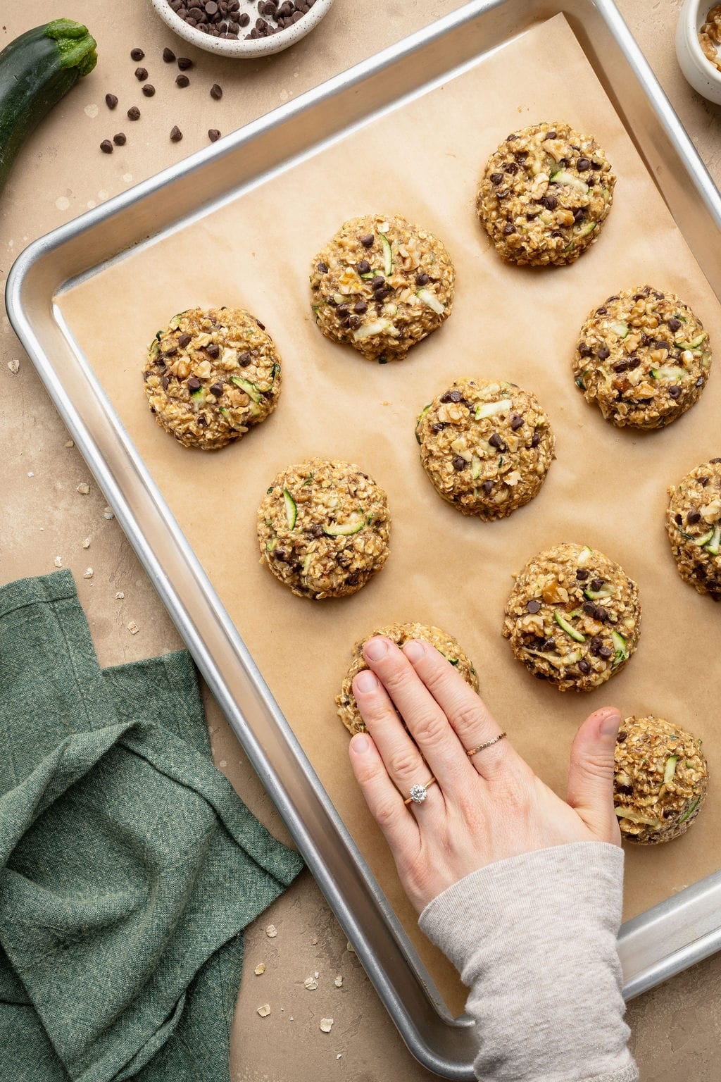 A hand flattening raw zucchini breakfast cookies on a parchment paper-lined baking sheet.