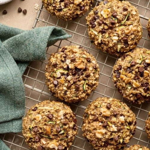Zucchini cookies on a wire rack, a blue kitchen towel and a small bowl of chocolate chips next to them.