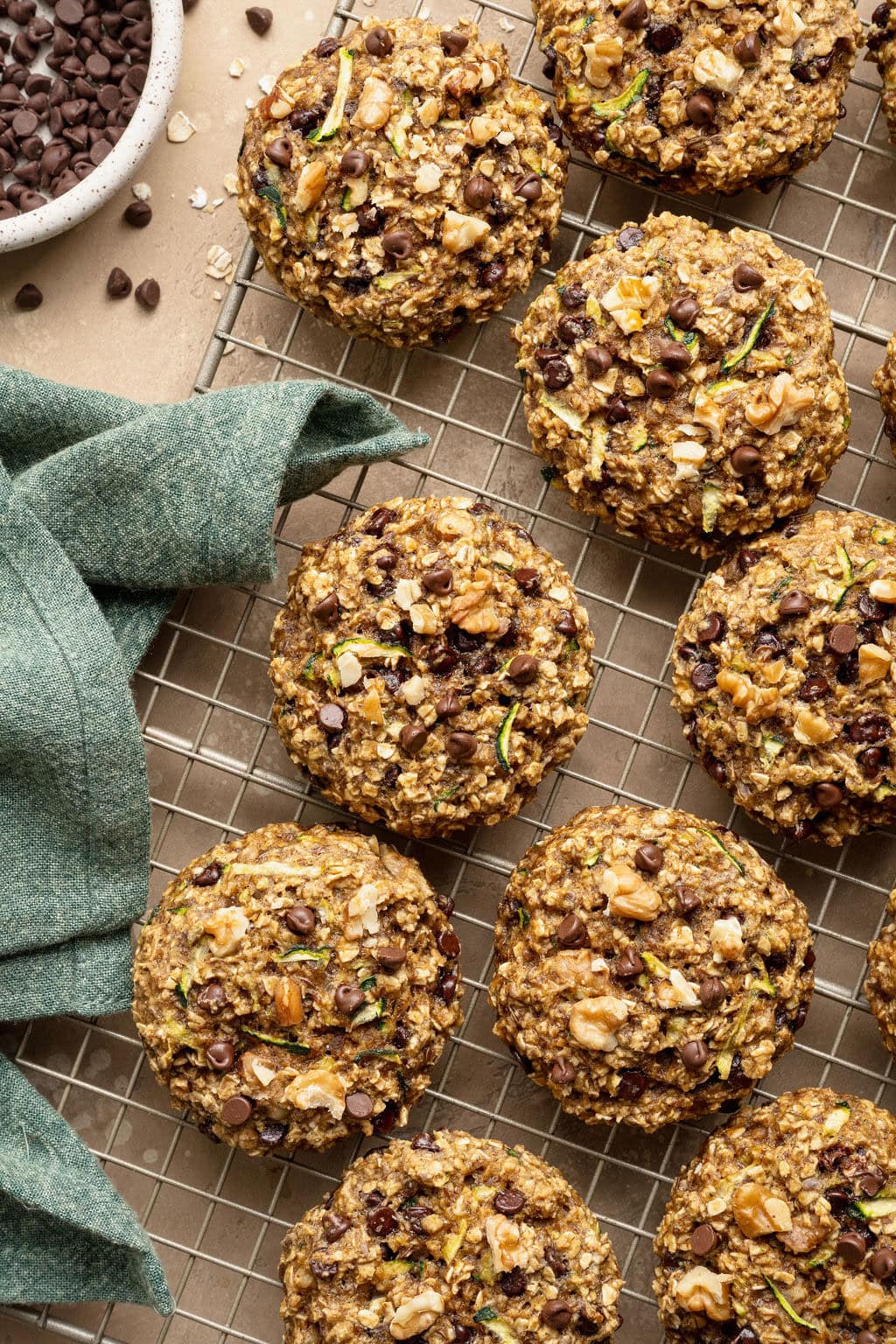 Zucchini cookies on a wire rack, a blue kitchen towel and a small bowl of chocolate chips next to them.