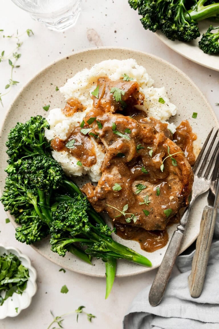A white plate with serving of smothered chicken over mashed potatoes with broccolini on the side