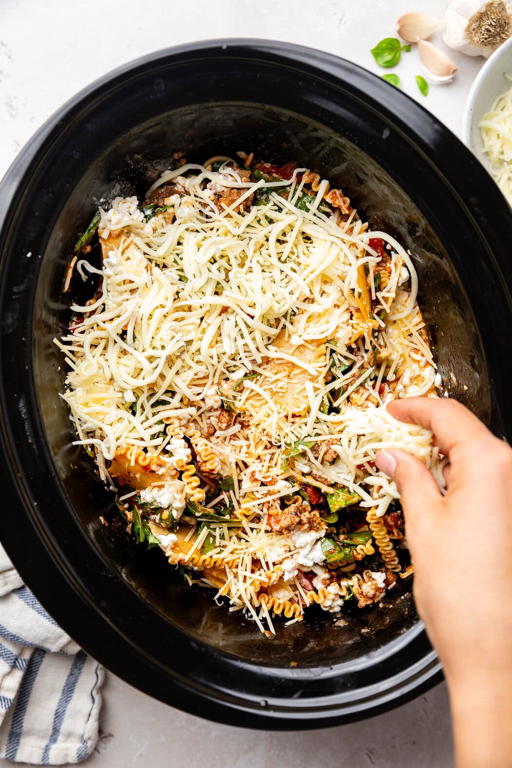 Overhead view of a slow cooker filled with ingredients for Crockpot Lasagna and a hand sprinkling cheese on top. 