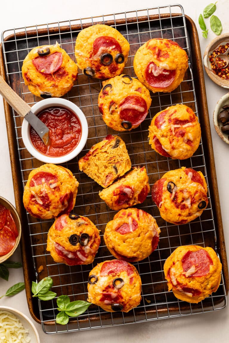 Overhead view of a cooling rack filled with easy pizza muffins filled with pepperoni pieces and black olives with a marinara sauce on the side. 