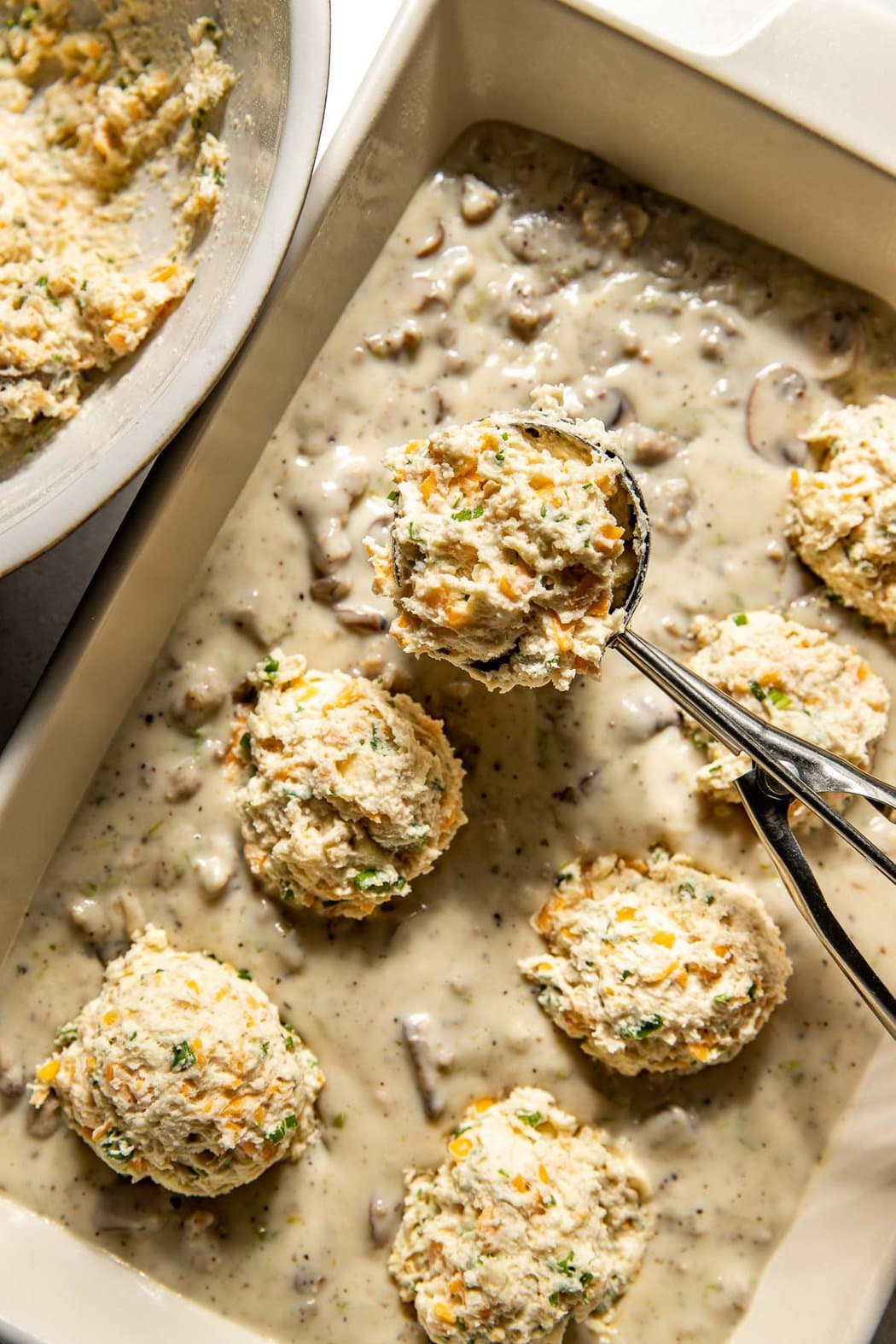 Overhead view of a pan of sausage gravy with a spoon scooping biscuit dough on top. 