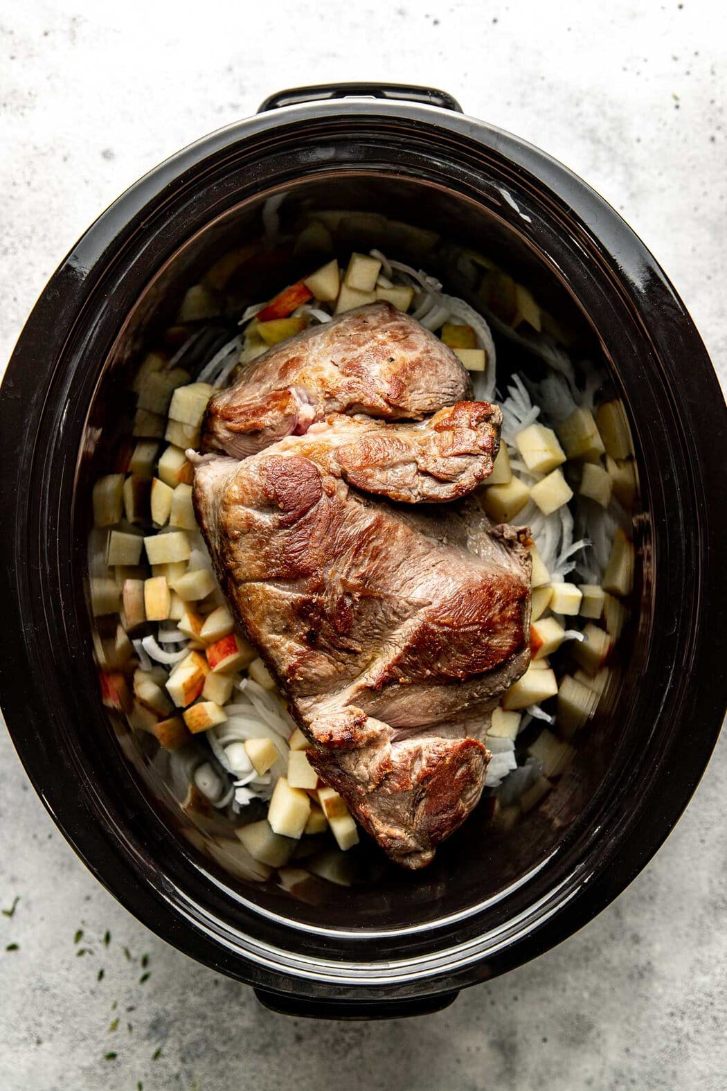 Overhead view of a crock pot filled with sliced onions, apple pieces, and a large, seared pork roast. 