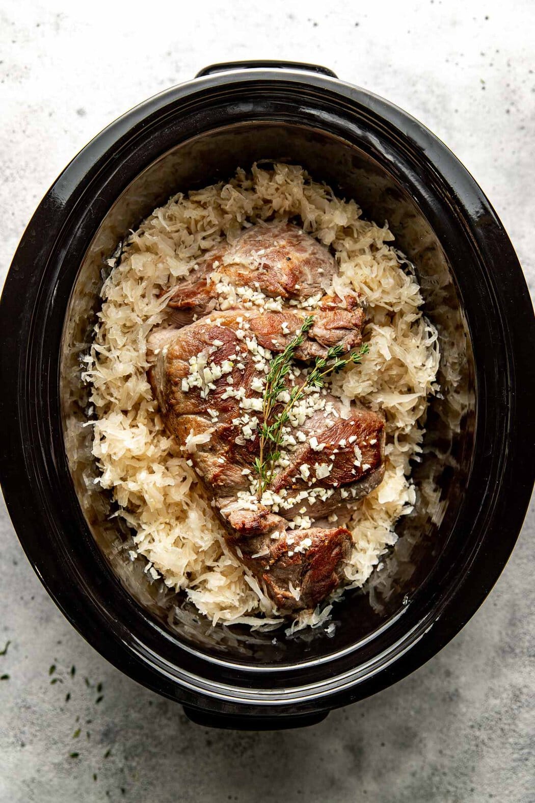 Overhead view of a crock pot filled with Pork and Sauerkraut topped with garlic and thyme. 