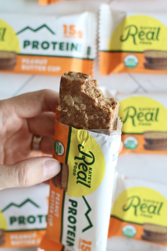 Close up view of a hand holding an opened kates real food bar with a bite out of the top to show the inside texture.