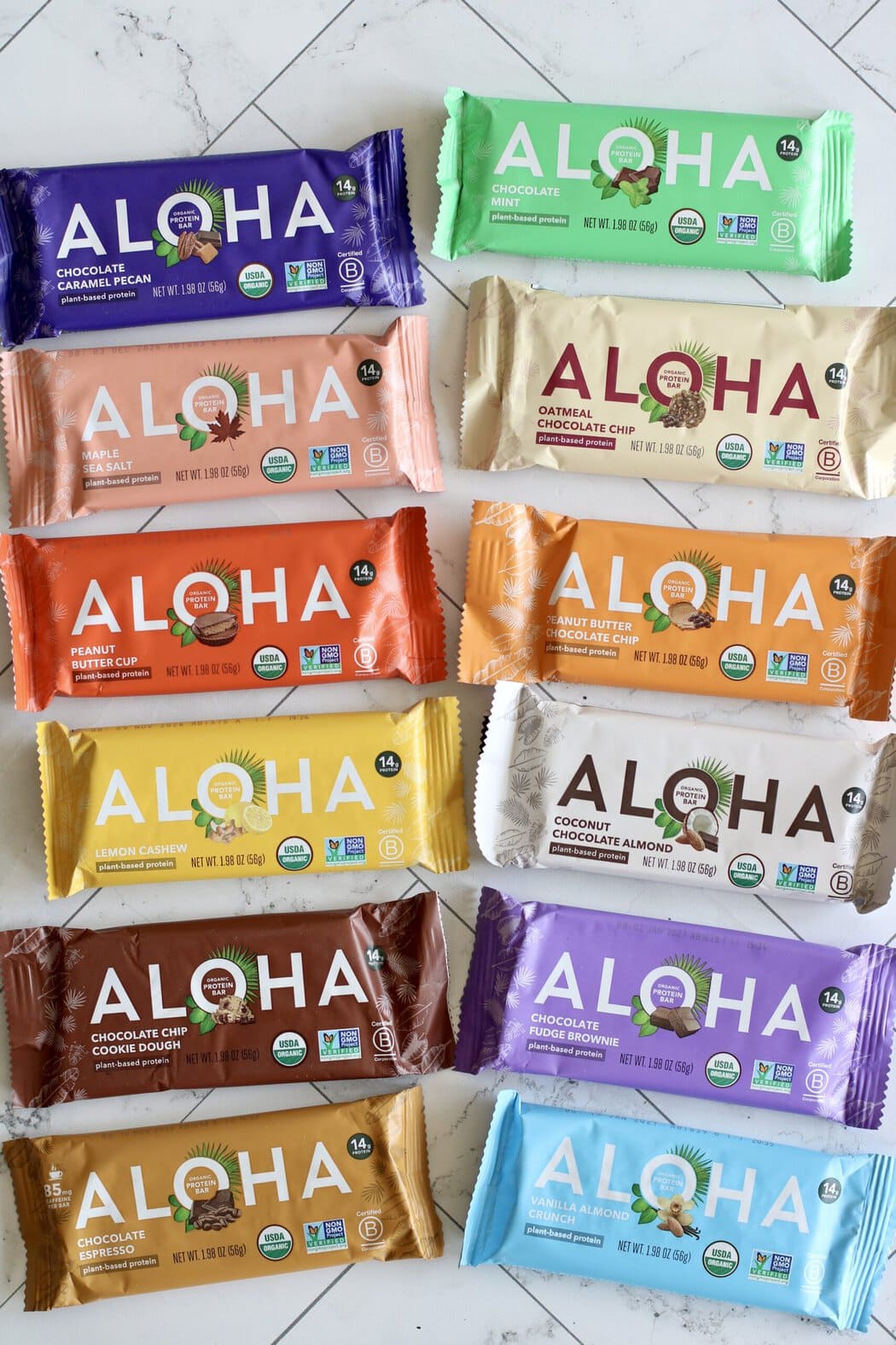 Overhead view of a variety of aloha brand protein bars arranged in lines on a white marble countertop.