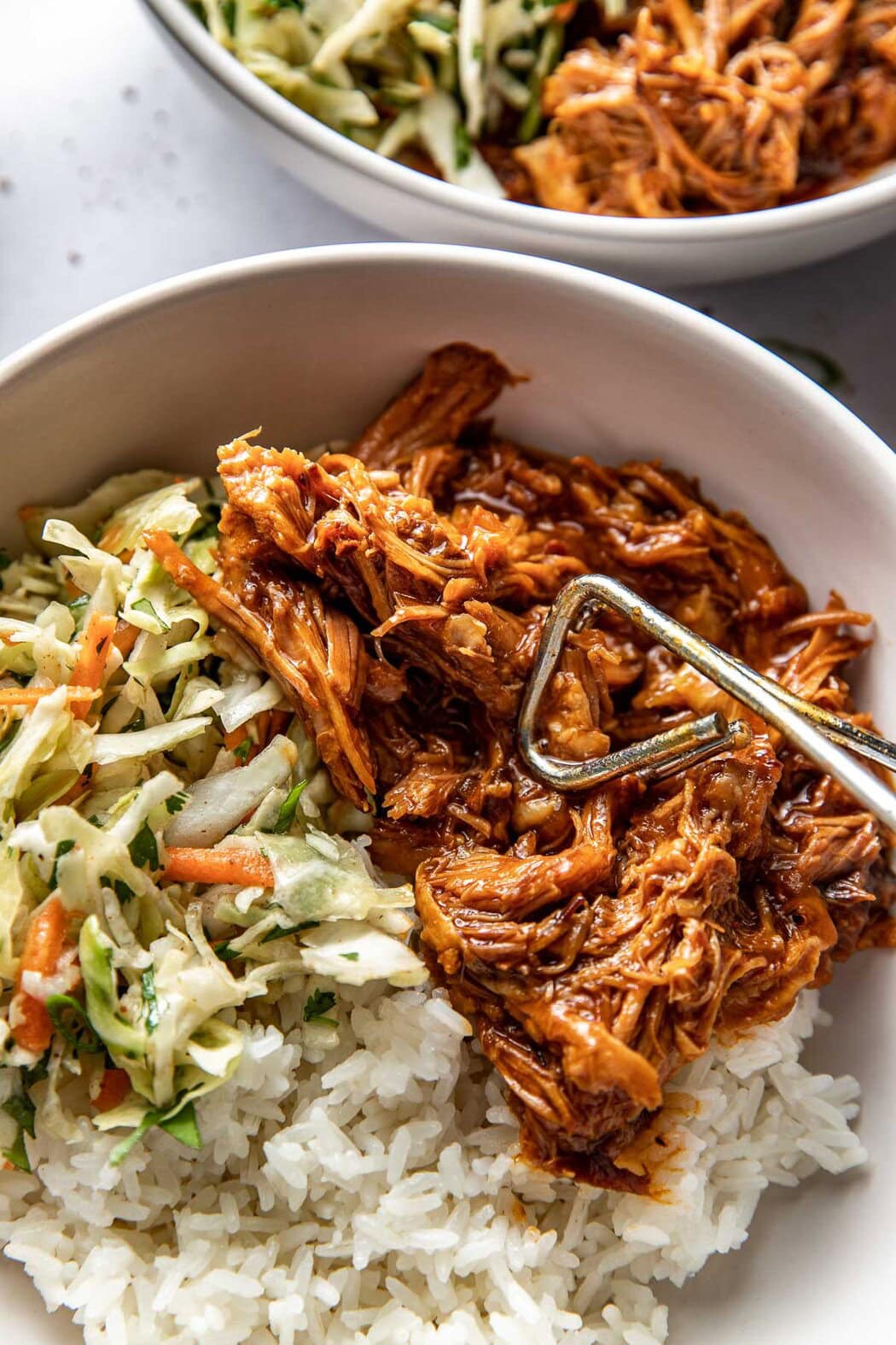 Close up view of a tongs nestling pulled pork into a bowl of rice and Cole slaw. 