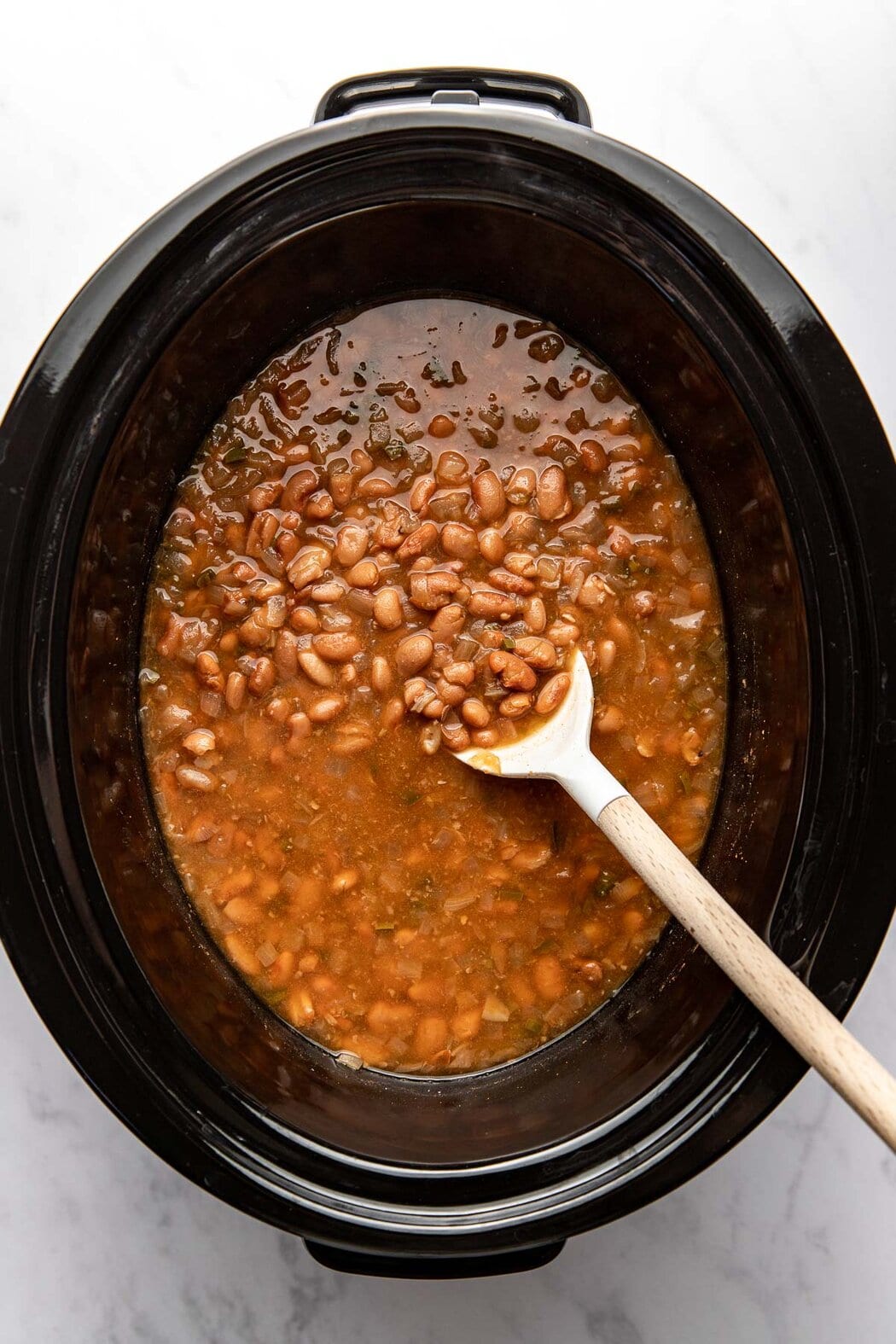 Overhead view of a slow cooker filled with refried bean ingredients being stirred with a wooden spoon.