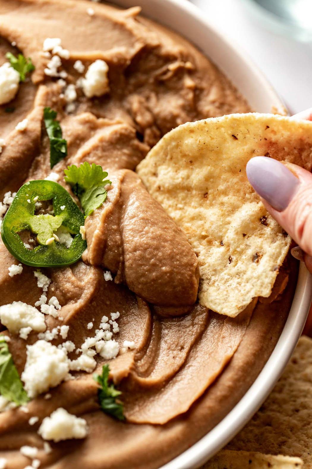 Close up view of a hand dipping a tortilla chip into refried beans topped with cheese and jalapeno.