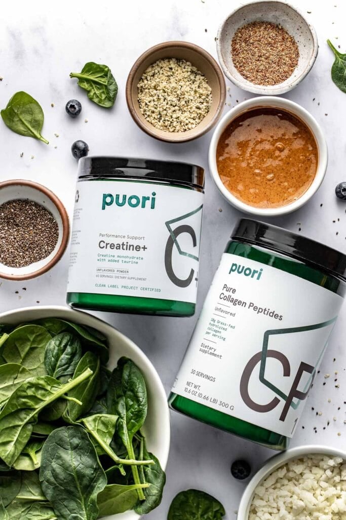 Overhead view of a canister of creatine and a canister of collagen next to small bowls of smoothie ingredients and a bowl of spinach