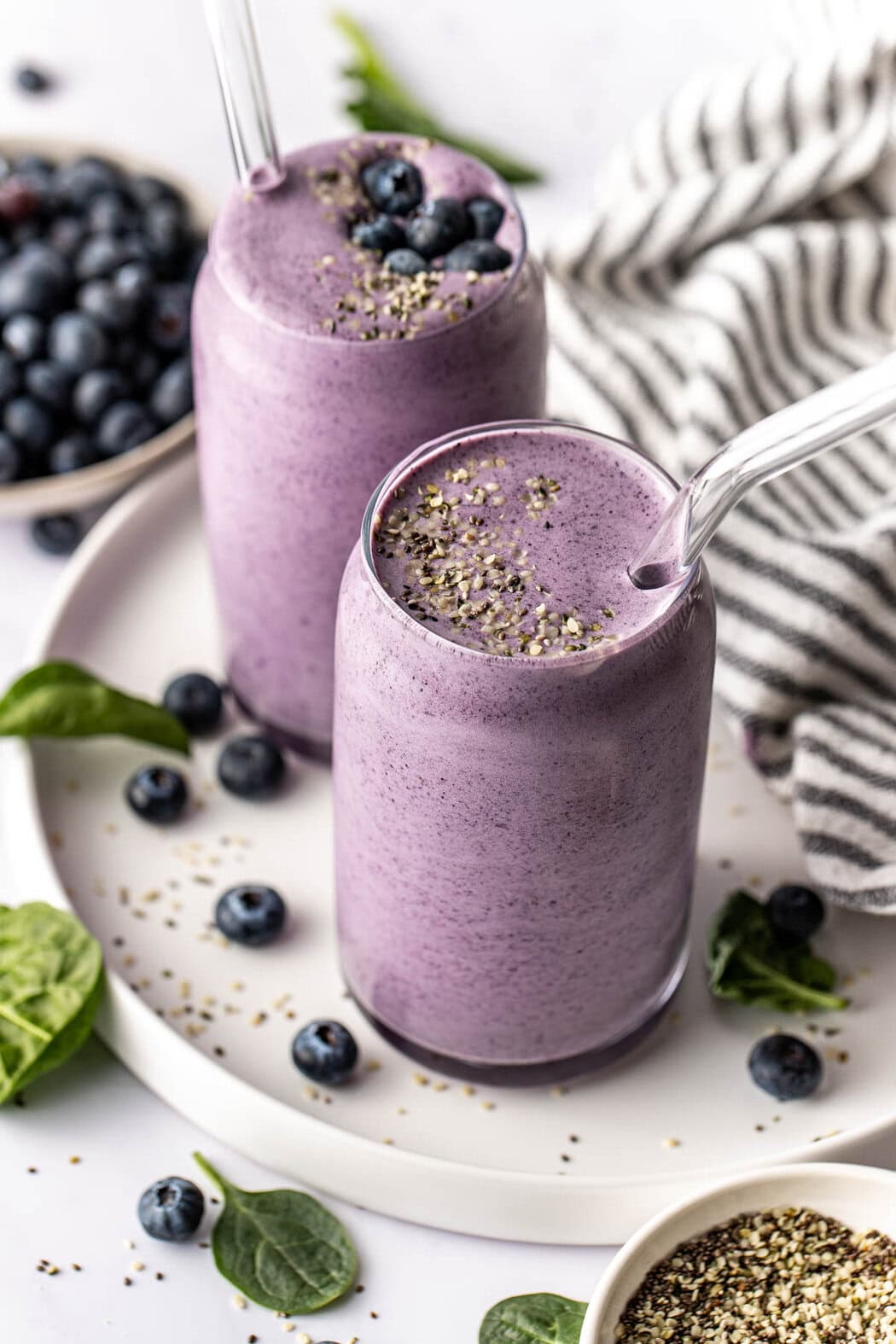 Close up view of two deep purple colored Post Workout Smoothies in glasses with glass straws topped with hemp seeds and blueberries.