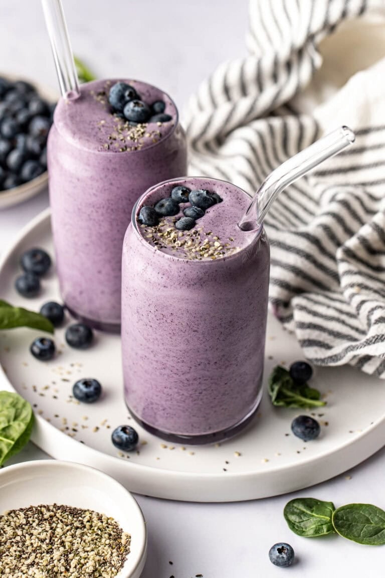 Overhead view of two colorful blueberry smoothies topped with fresh blueberries and hemp hearts in glasses with glass straws. 