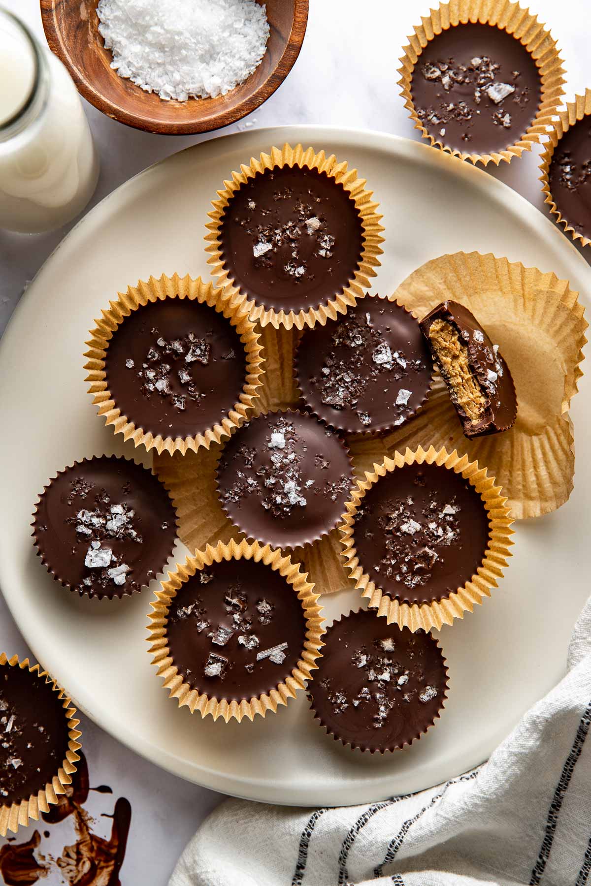 Overhead view of a plate filled with Peanut Butter Protein Cups and tan paper liners topped with flakey sea salt. 
