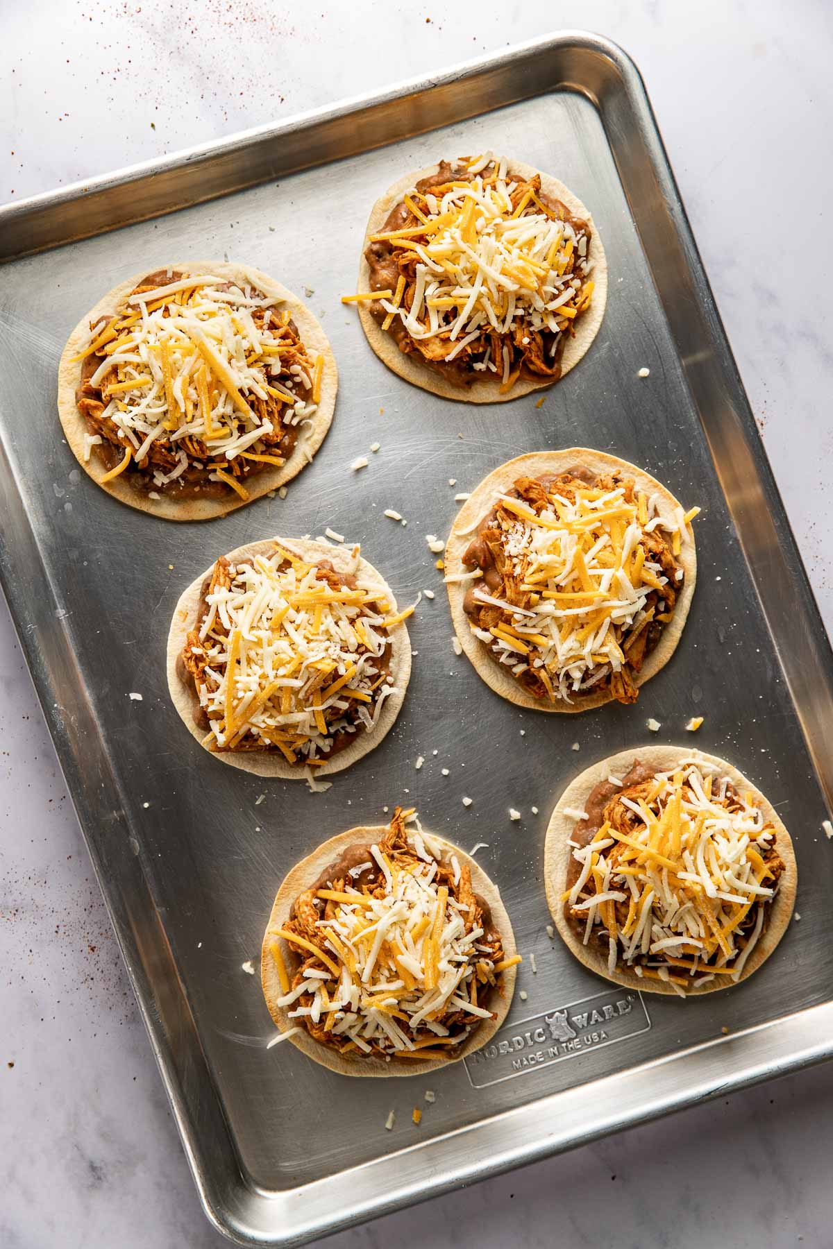 Overhead view of a pan of Sheet Pan Chicken Tostadas ready for the oven topped with shredded cheese. 