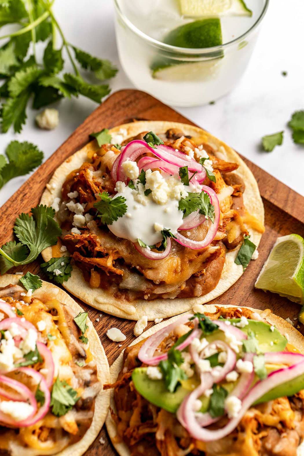 Close up view of Sheet Pan Chicken Tostadas on a wooden cutting board topped with melted cheese, sour cream, and fresh cilantro.