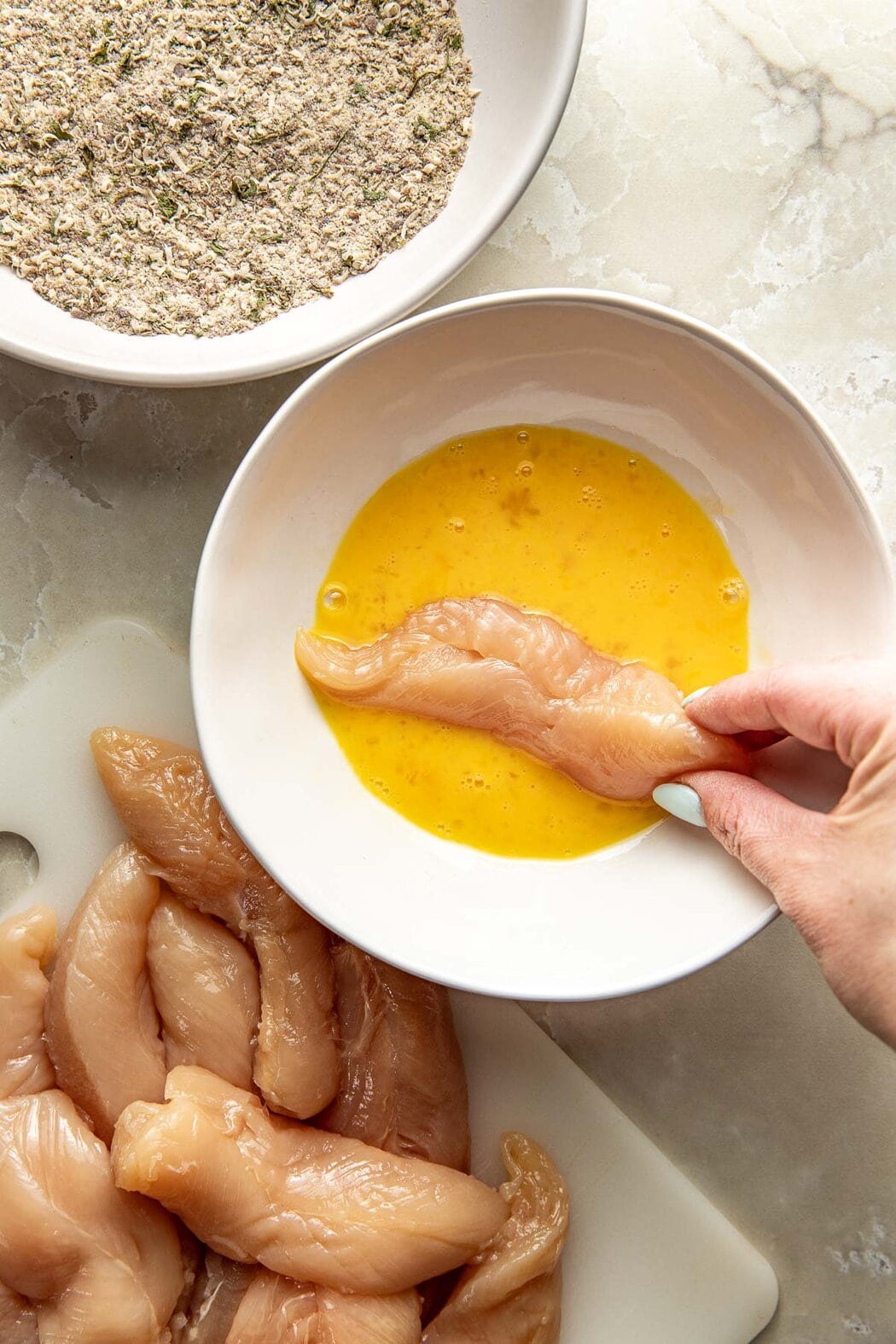 Overhead view of a hand dipping a raw chicken tender into an egg wash.