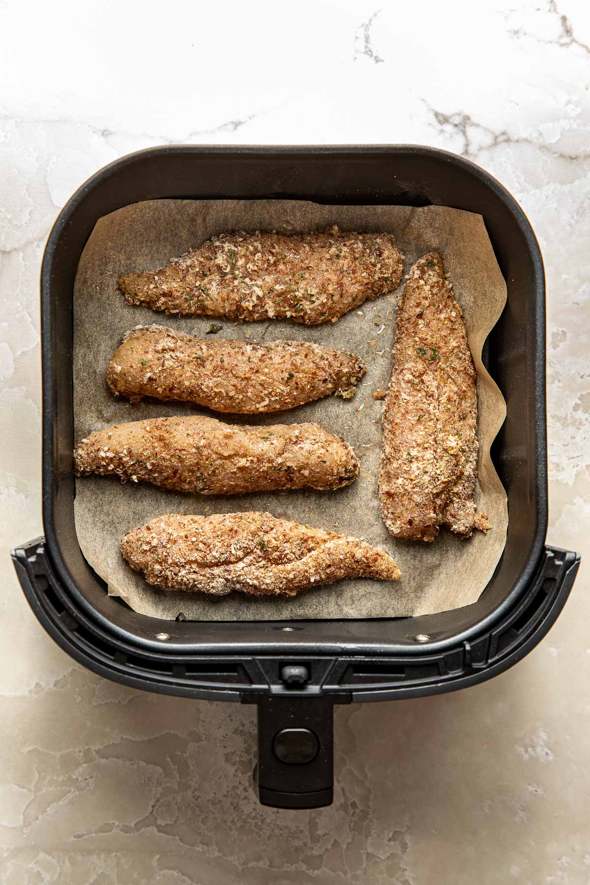 Overhead view of an air fryer basket lined with parchment paper and filled with breaded chicken tenders ready for baking
