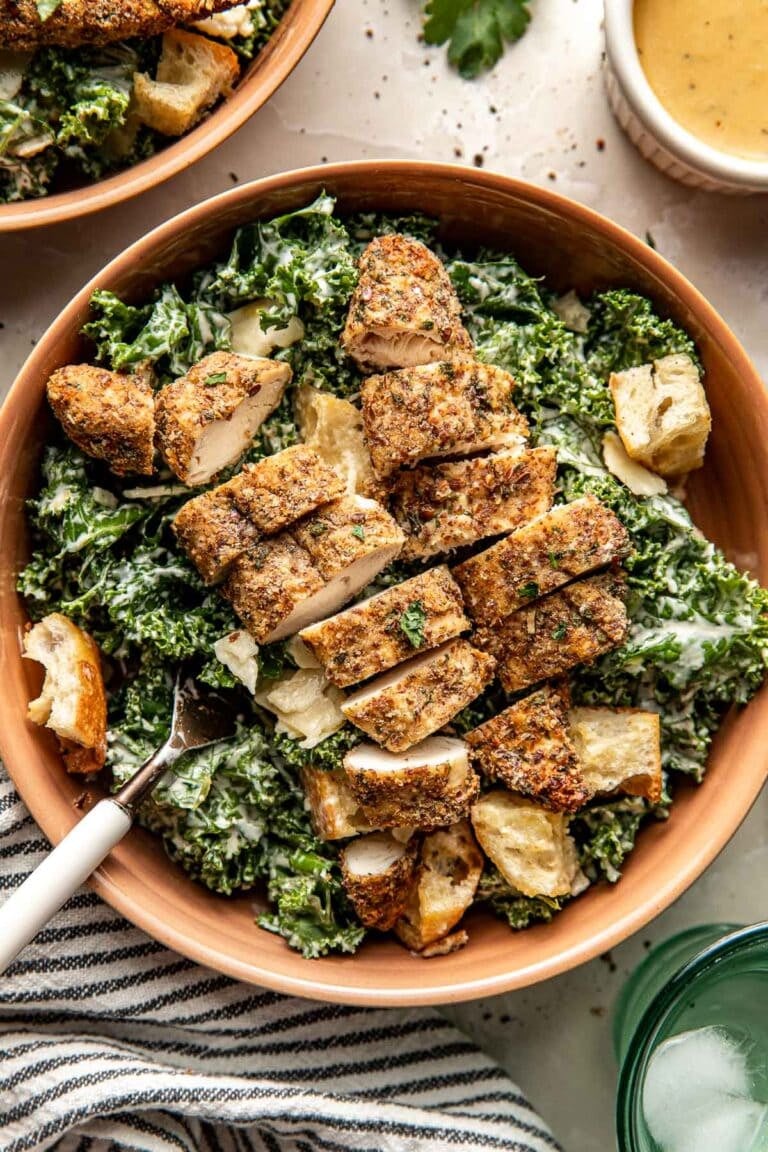 Overhead view of a bowl of kale salad topped with sliced chicken tenders topped with fresh herbs and black pepper.