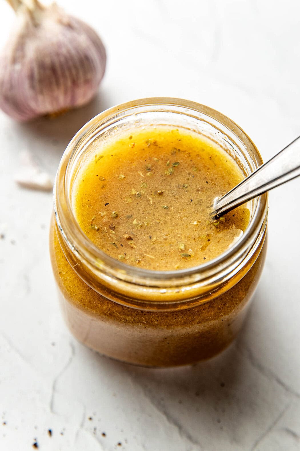 Close up view of a glass jar of Red Wine Vinaigrette with a spoon stirring the mixture to show the spices and herbs. 