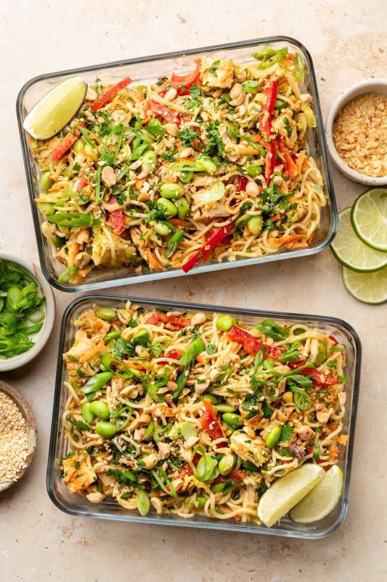 Overhead view of two glass meal prep containers filled with cold noodle salad with peanut sauce topped with fresh herbs and edamame beans.