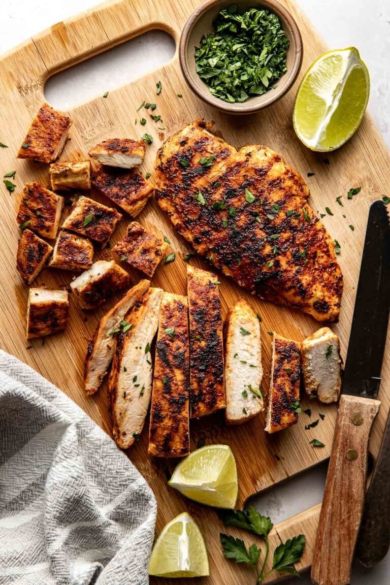 Overhead view of a wooden cutting board filled with blackened chicken breasts sliced next to lime wedges.