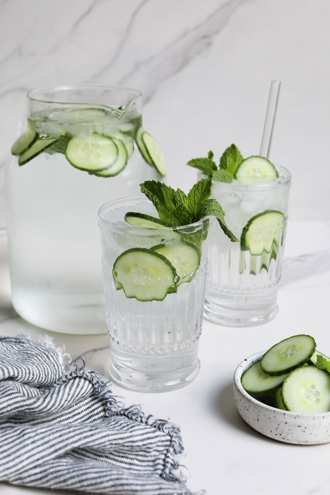 Close up view of a glass water pitcher and two glasses filled with cucumber water and topped with mint leaves. 