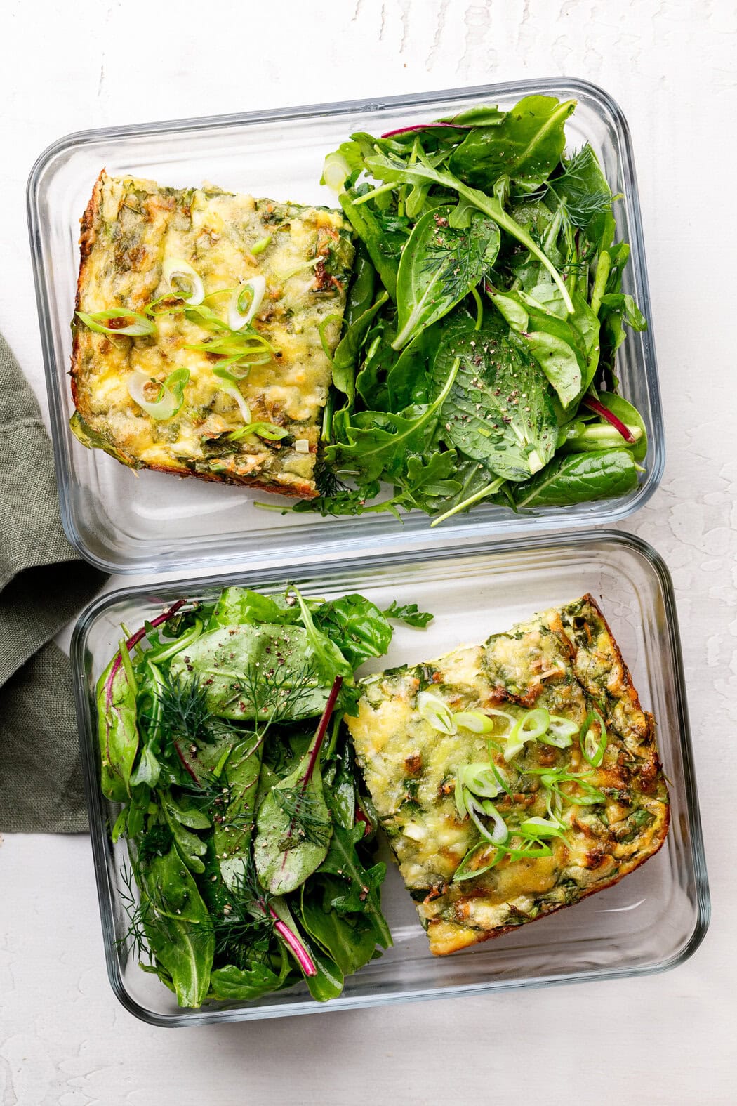 Overhead view of two glass meal prep containers filled with Cottage Cheese Egg Bake portions and mixed greens on the other side.
