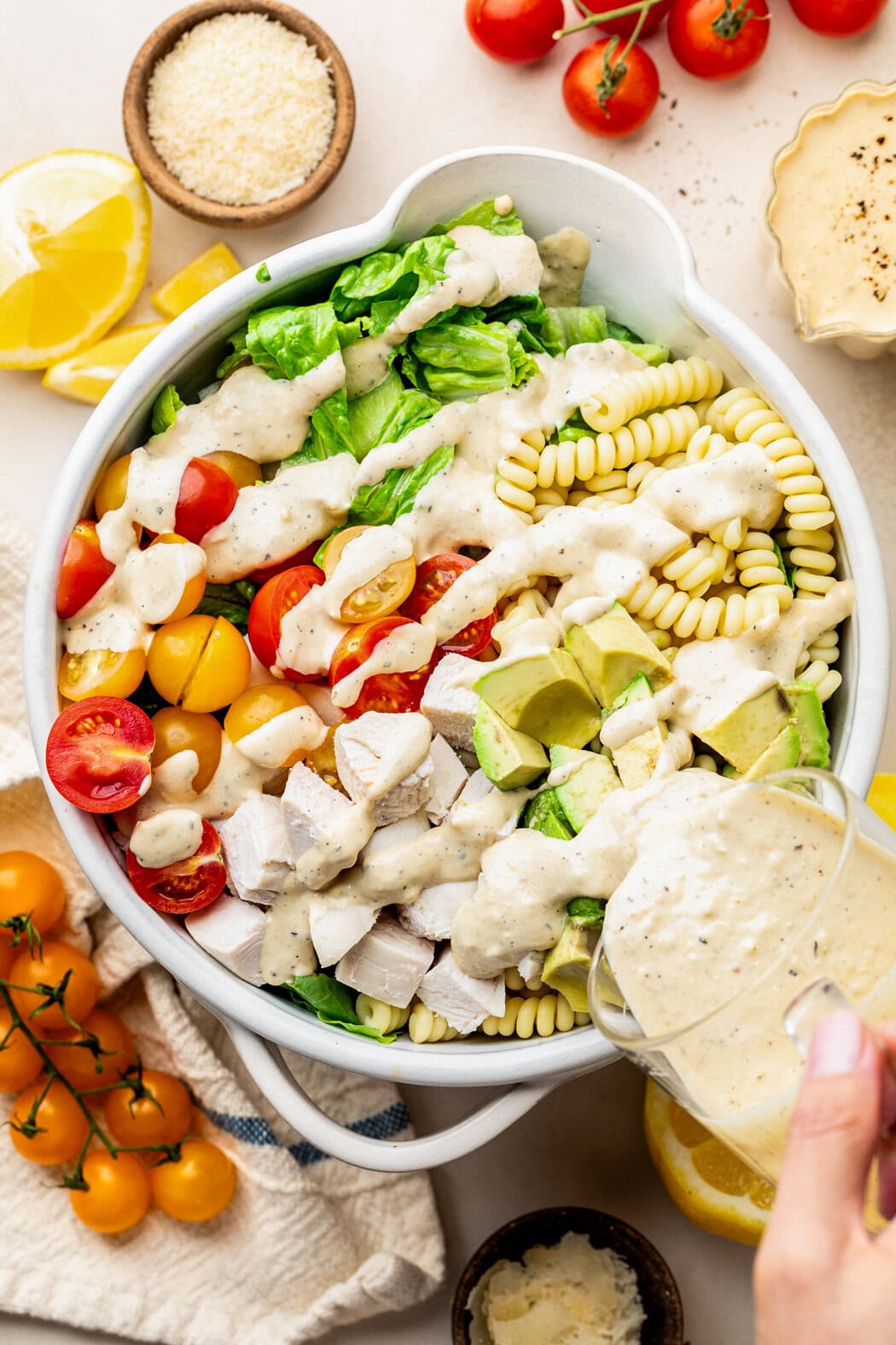 Overhead view of a bowl of Chicken Caesar Pasta Salad with a hand pouring the Caesar sauce over the top of the ingredients. 