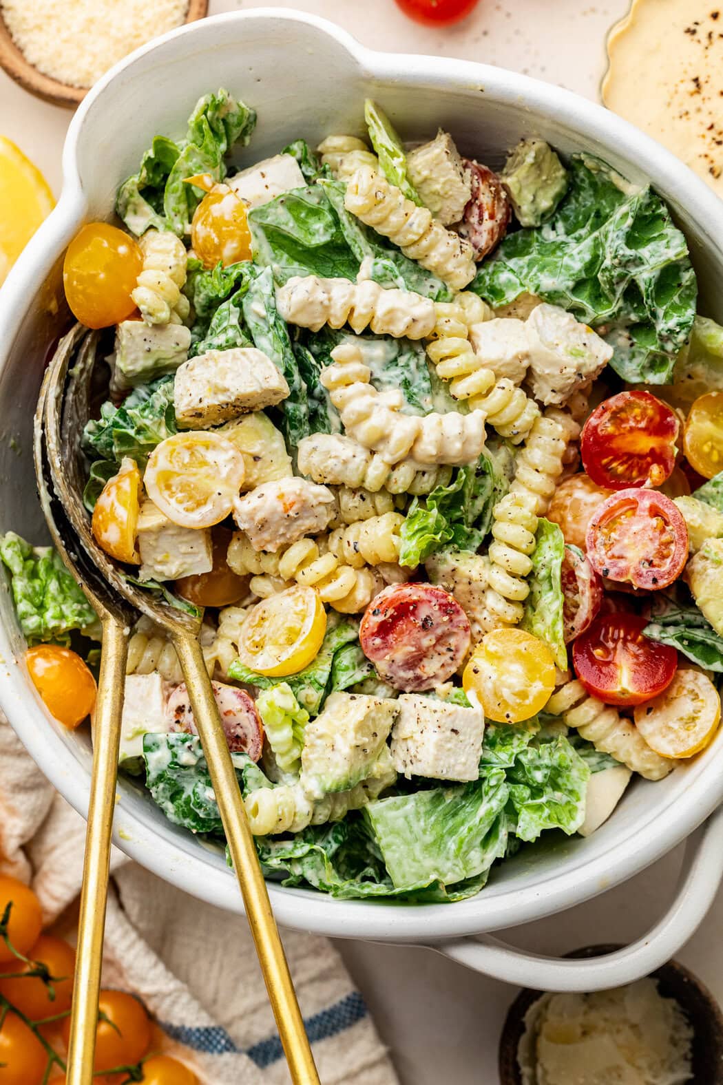 Overhead view of a large white bowl filled with freshly tossed Chicken Caesar Pasta Salad with two serving spoons in the bowl. 